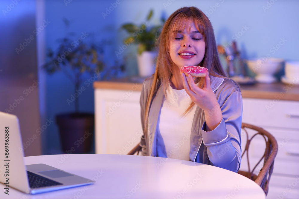 Young woman with tasty doughnut in kitchen at night