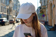 © vejaa - Teenage girl in white t-shirt and blank white baseball cap looking to her phone while standing in European old town street. White cap mockup template