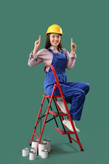 Wall Mural - female asian decorator on stepladder pointing at something against green background