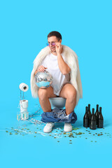 Wall Mural - Young man with disco ball sitting on toilet bowl during party against blue background