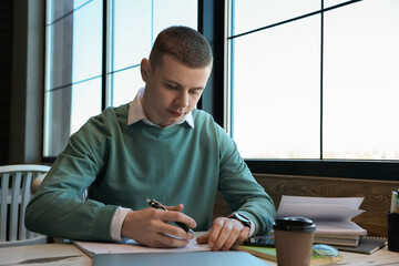 Wall Mural - Young male student with notebooks studying at table in cafe