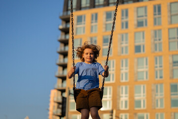 Wall Mural - Excited kid swinging on chain swing on city kids playground. Swing ride. Cute child having fun on a swing on summer sky background. Blonde little boy swings at kid playground. Child swinging high.