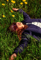 Wall Mural - Happy relaxing kid girl with beautiful long curly hair smiling lying on the green grass with yellow flowers on summer bright sunny day background in fashion violet sunglasses. Lifestyle