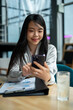 © bongkarn - A beautiful Asian businesswoman or female boss is checking her messages on her smartphone at a desk.