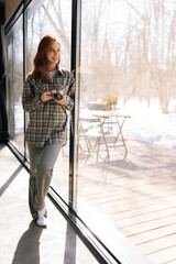 full length vertical portrait of happy young woman enjoying drinking cup of coffee at home standing 