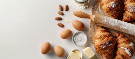Poster - Aerial view of fresh croissants on a wooden board with dough, rolling pin, butter, and eggs on a white backdrop allowing for a copy space image.