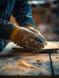 © Boontharika - Close-up of a craftsman sanding wood, wearing gloves. Dust particles flying, focused shot of hands woodworking in a workshop.