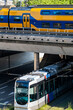 © yaqui_villegas - A yellow and blue train crosses an elevated overpass while a city tram navigates below on the street level, showcasing a multi-layered urban transport system.