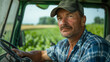 © ifoto - A farmer in his mid-50s, wearing a blue plaid shirt and green baseball cap with a gray brim, sits behind the wheel of an open tractor cab looking out at crops