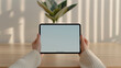 © Iana - woman's hands holding black tablet iPad pro horizontal with white blank screen, sitting at a simple wooden desk, with only a small potted plant in the background, minimalism