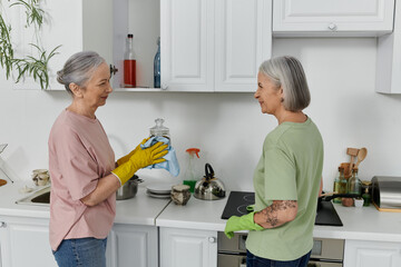 A mature lesbian couple cleans their modern apartment kitchen together. One woman is wearing yellow gloves and wiping a glass jar.