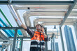 © Washburn - A construction engineer in a safety vest and helmet inspects industrial pipes on a rooftop while communicating via walkie-talkie.