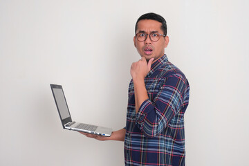 Wall Mural - Side view of a man showing confused face at the camera while holding a laptop
