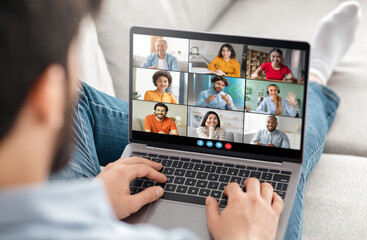 Poster - A man is sitting on a couch at home, using a laptop to participate in a video conference call with several other people. He is typing on the keyboard and appears to be actively engaged