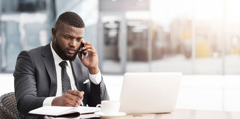 Wall Mural - African Business Guy Multitasking Talking On Cellphone Working On Laptop And Taking Notes Sitting In Outdoor Cafe. Busy Lifestyle Concept