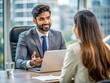 © Man888 - Indian businessman conversing with female interviewer, gesturing while seated at modern office desk with laptop, showcasing confident and professional demeanor.