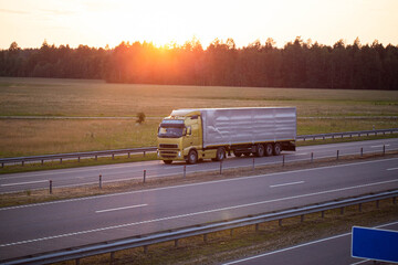 A truck driver on a tractor with a tilt semi-trailer transports cargo against the backdrop of a forest and sunset in the evening in summer. The concept of the work 