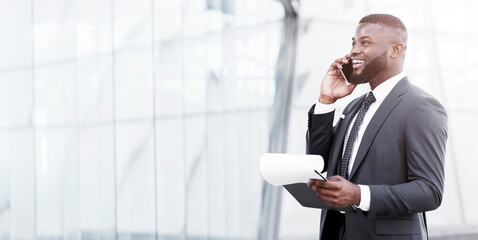 Wall Mural - Business Calls Concept. African American Business Guy Talking On Phone Holding Papers Negotiating Deal Standing In City. Copy Space