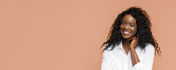 Wall Mural - This image shows a young African American woman with curly black hair smiling at the camera. She is wearing a white button-down shirt and is standing against a peach-colored background.