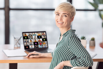 Poster - Mature woman with short blonde hair sits at a desk in an office setting with a laptop open in front of her, looking at the camera and smiling, with her right hand resting on the back of her chair