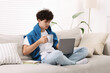 © New Africa - Teenager holding cup of drink working with laptop on sofa at home. Remote job
