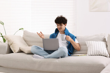 Sticker - Teenager holding cup of drink working with laptop on sofa at home. Remote job