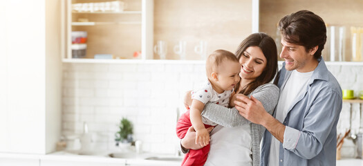 Wall Mural - A happy family stands in a modern kitchen, with the mother holding their baby while the father smiles and looks at the child.