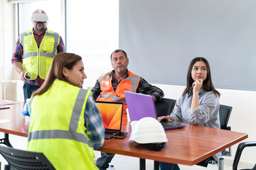 A group of construction workers are sitting around a table having a meeting about virtual reality. They are wearing , hard hats and safety vests. There are two men and two women in the group.