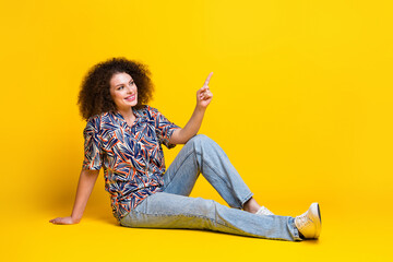 Wall Mural - Full size photo of pretty young girl sit floor relax look point empty space wear trendy colorful outfit isolated on yellow color background