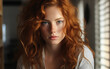 © imagineRbc - A close-up portrait of a young woman with long, curly red hair. She is looking directly at the camera with a serious expression. The photo was taken indoors