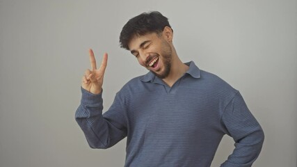 Canvas Print - Young arab man standing over isolated white background, smiling happily and winking, showing victory sign with two fingers 'v' and radiating confidence, symbolizing success and win