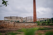 © Raivo - limbazi, Latvia, June 29, 2024 - Abandoned, partially demolished building with a tall red chimney against a cloudy sky, surrounded by rubble and overgrown vegetation...
