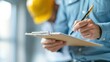 © KanitChurem - Close-up of a construction worker's hands writing on a clipboard.  The worker is wearing a blue shirt.  A yellow hard hat can be seen in the background.