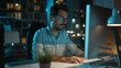 © VK Studio - Man diligently working on a computer in a contemporary office with city lights twinkling through the window at night.