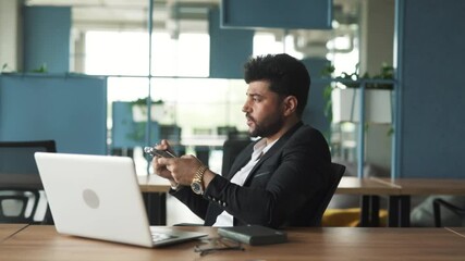portrait of a handsome, stylish young man of Arab descent sitting in a modern business center office with a laptop, recording a voice message on his smartphone while speaking