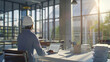 © VK Studio - A construction worker in a hard hat, viewed from behind, works on his laptop in a sunlit modern office with floor-to-ceiling windows.