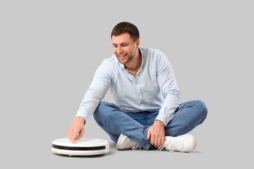 Wall Mural - Handsome man with robot vacuum cleaner sitting on light background