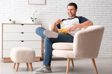 Sticker - Handsome happy young man with bowl of tasty potato chips sitting on sofa at home