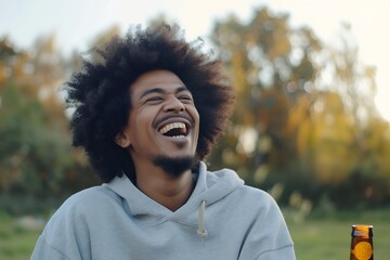 Wall Mural - Portrait of happy young man with afro hair wearing grey sweatshirt drinking beer bottle and laughing outdoor, real photo 