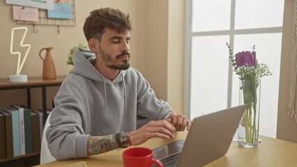 Wall Mural - Handsome young man with beard and tattoos feeling stressed while working on laptop in a modern home interior.