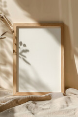 A blank white poster mockup in an oak frame, resting on a beige cloth background with soft shadows and natural lighting