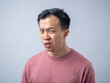 © Daniel - An Asian man in a pink shirt makes a skeptical and uncomfortable expression, as if reacting to something unpleasant. The plain light blue background highlights his displeased