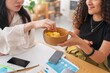 © Studio Marmellata - Two women sharing snacks while working at a desk in a modern office Focus on their casual interaction