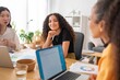 © Studio Marmellata - Young woman staring at camera sitting at a table in a modern office space, with laptops and documents visible
