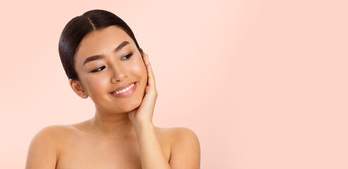 Wall Mural - A young Asian woman with dark hair pulled back smiles and looks at copy space while touching her face. She is wearing no makeup and has a soft, natural look. The background is a light pink color.