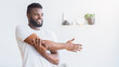 © Prostock-studio - A young black man with short dark hair and a beard is stretching his arm in front of him while standing in a white room, copy space