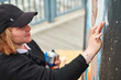 © TRAVELARIUM - Female artist in black cap is painting picture with paint spray can spraying it onto canvas at outdoor street exhibition, side view of female art maker