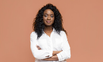 Wall Mural - African American woman with long, curly hair wearing a white button-down shirt with her arms crossed. She is standing against a peach-colored background. She has a confident expression on her face.