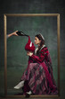 © master1305 - Portrait of young medieval royal person, woman sitting inside golden frame with giant wine glass against vintage green background. Concept of history, comparison of eras, remake restaurant