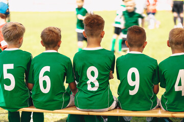 Wall Mural - Group Of School Boys in Sports Team. Kids Sitting On Wooden Bench. Children in Green Sports Uniforms With White Numbers on their Backs. Youth Soccer Team Compete in a Tournament Match